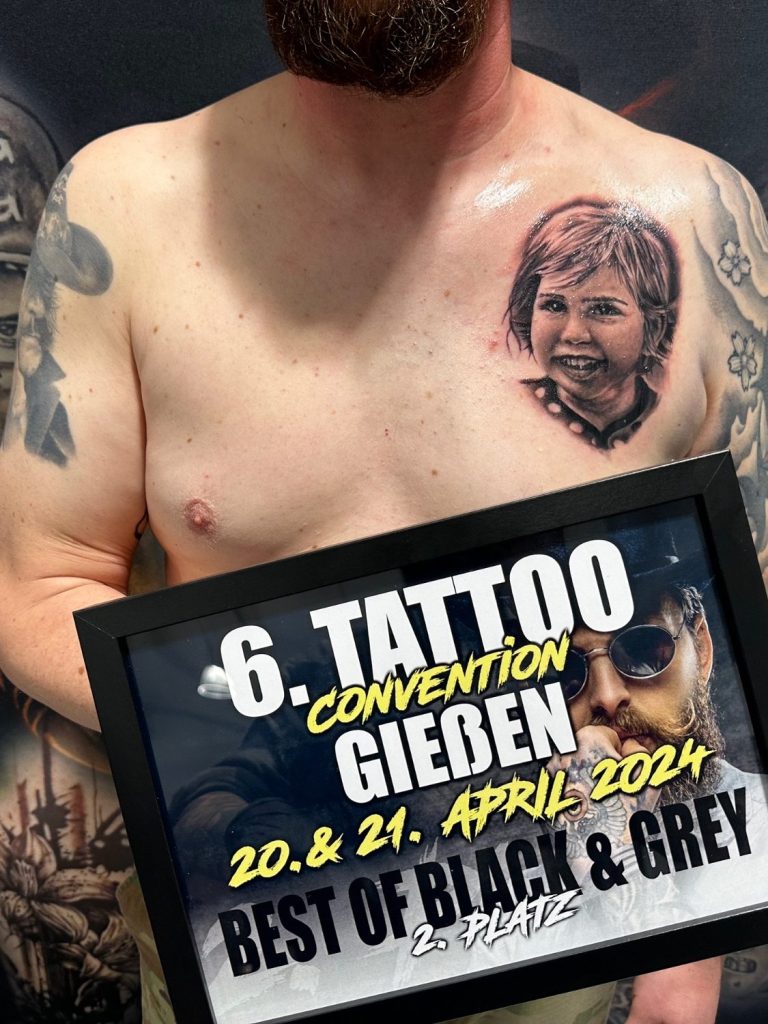 Ein Mann mit tätowierter Kinderporträt auf der Brust hält einen Preis der Tattoo-Convention in Gießen.marburg tattoo, tattoo marburg, tattoo